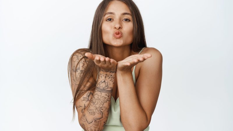 Flirty feminine woman with tattoo, sending air kiss on open hands, pucker lips cute, standing over white background.